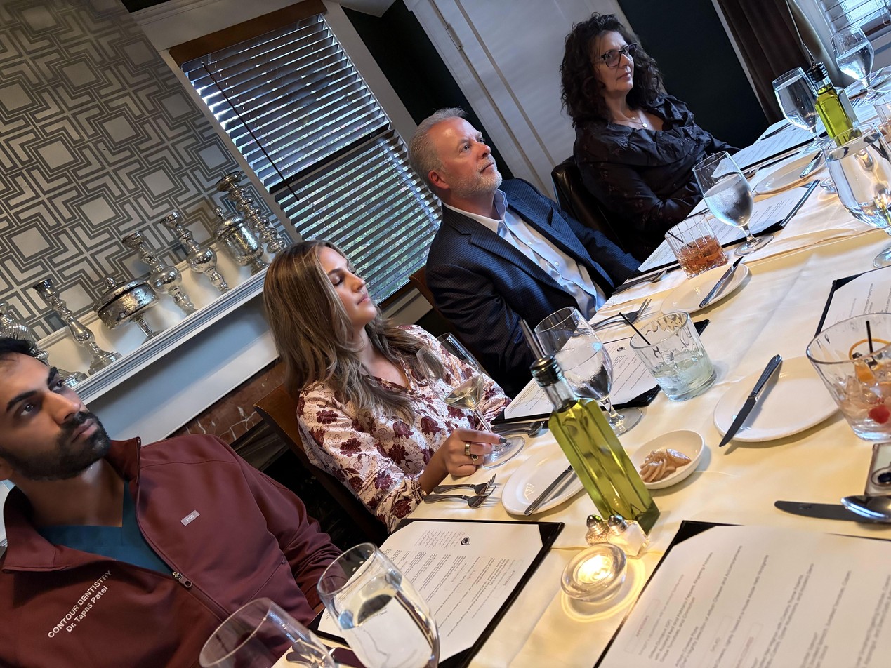 Four people seated along a dinner table set with menus, wine and water glasses, and olive oil, in front of a window with blinds and a mantel of silver candlesticks at The Fig Tree in Charlotte | The Paubox Table in Charlotte