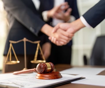 Image of two people shaking hands, near a desk that holds a scale and gavel. 