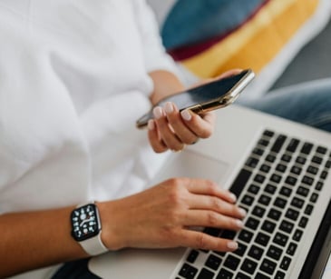 Image of someone typing on their computer while holding a phone. 