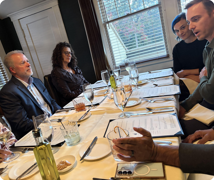 People talking around a dinner table set with water glasses, olive oil, and menus beside a window with blinds at The Fig Tree in Charlotte | The Paubox Table in Charlotte