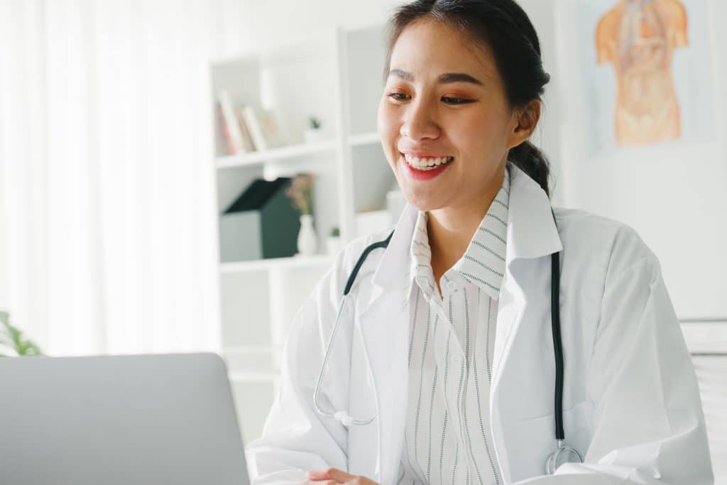 Young Asia lady doctor in white medical uniform with stethoscope using computer laptop talking video conference call with patient at desk in health clinic or hospital. Consulting and therapy concept.