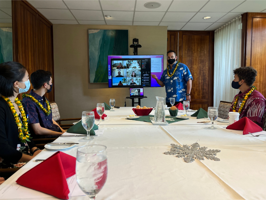 Masked people in a dining hall meeting with a monitor showing a Zoom meeting with a speaker near the monitor